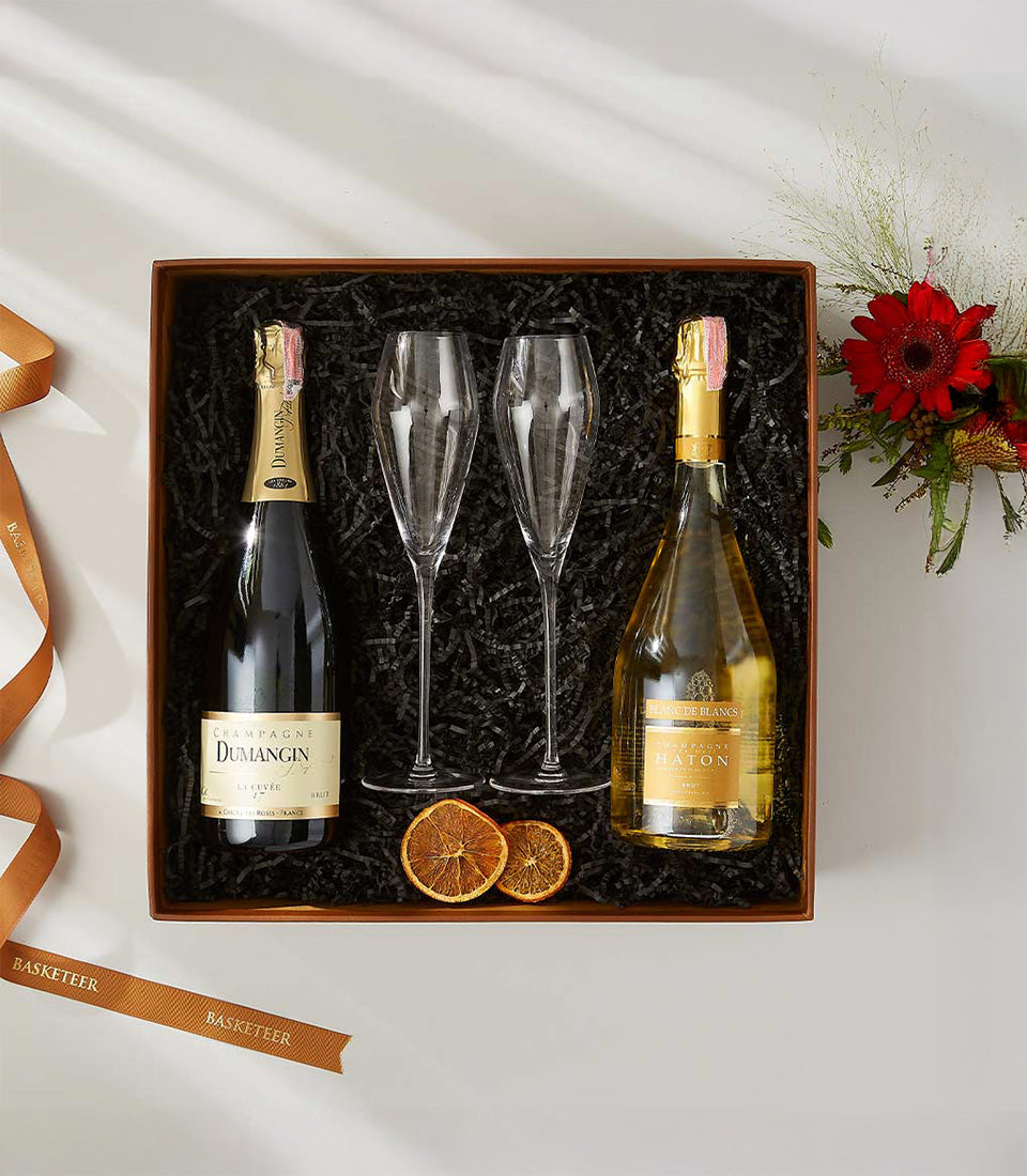Luxury champagne gift box with two bottles, flutes, and dried citrus on black shredded paper, tied with gold ribbon