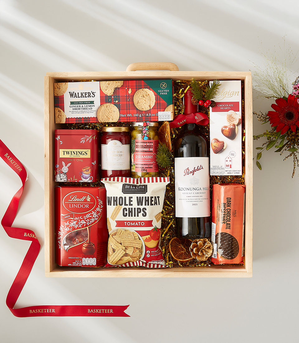 Wooden Christmas hamper with red wine, shortbread, tea, jam, and gourmet treats with gold tinsel and red ribbon