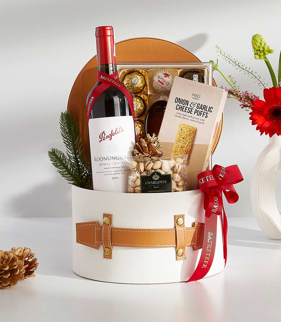 Luxury red wine gift hamper with chocolates, gourmet snacks, and festive holiday greenery in white cylindrical box