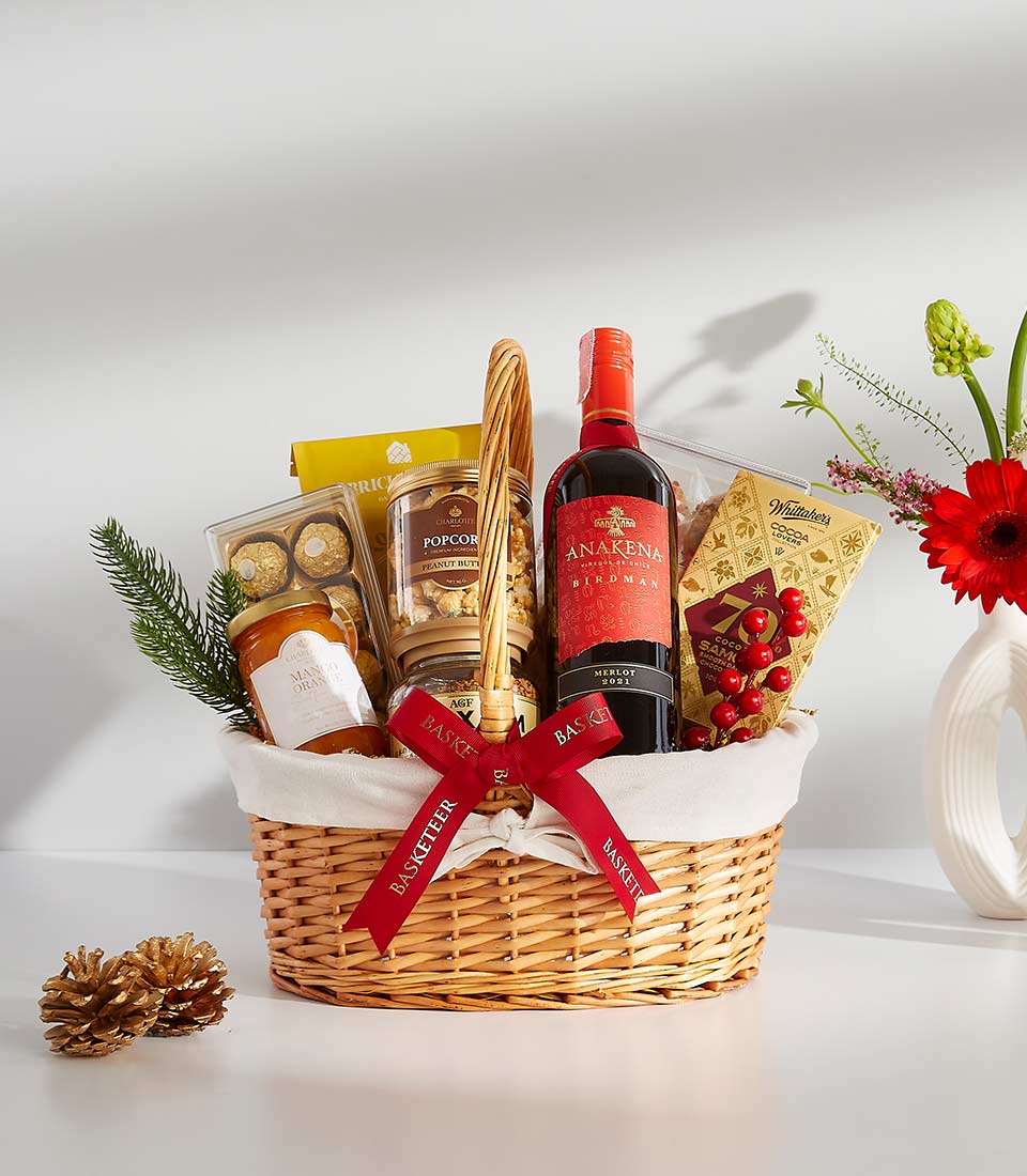 Woven wicker gift basket with red wine, gourmet snacks, chocolates, and fresh red gerbera daisies