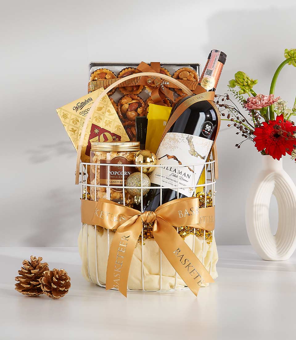 Festive white wire gift basket with red wine, gourmet snacks, and gold satin ribbon bow