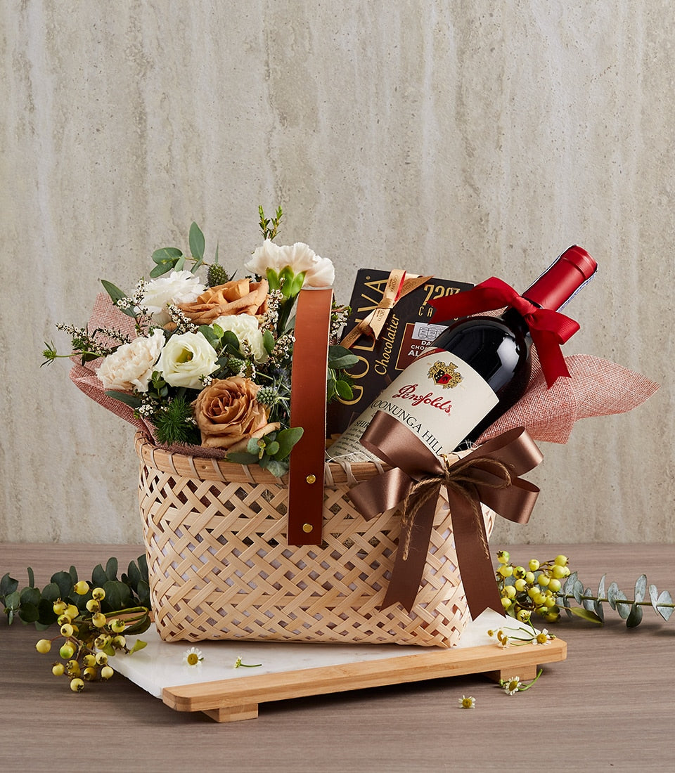Woven bamboo gift basket with red wine, white and peach roses, and chocolate box for anniversary celebrations