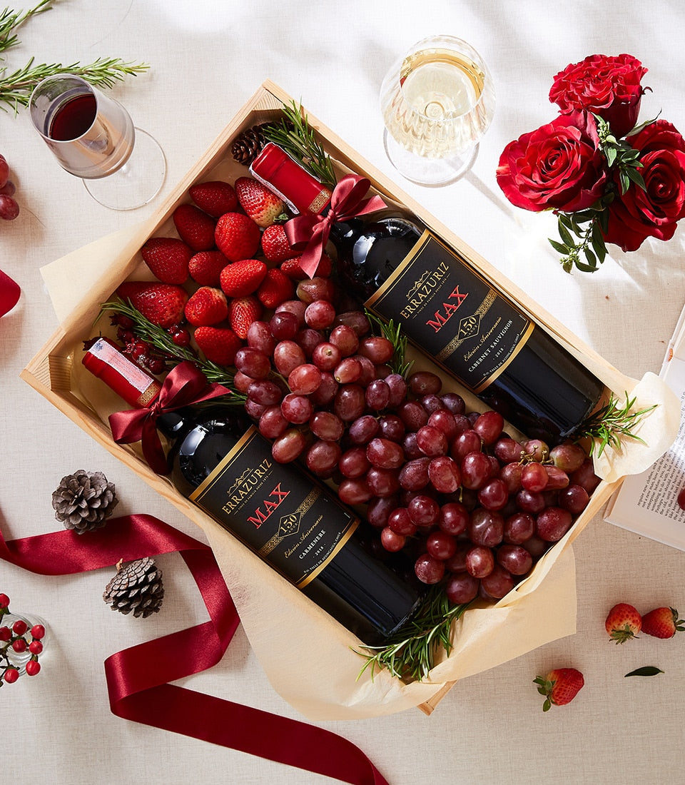 Wooden gift box with two red wine bottles, fresh red grapes, strawberries, rosemary, and pinecones