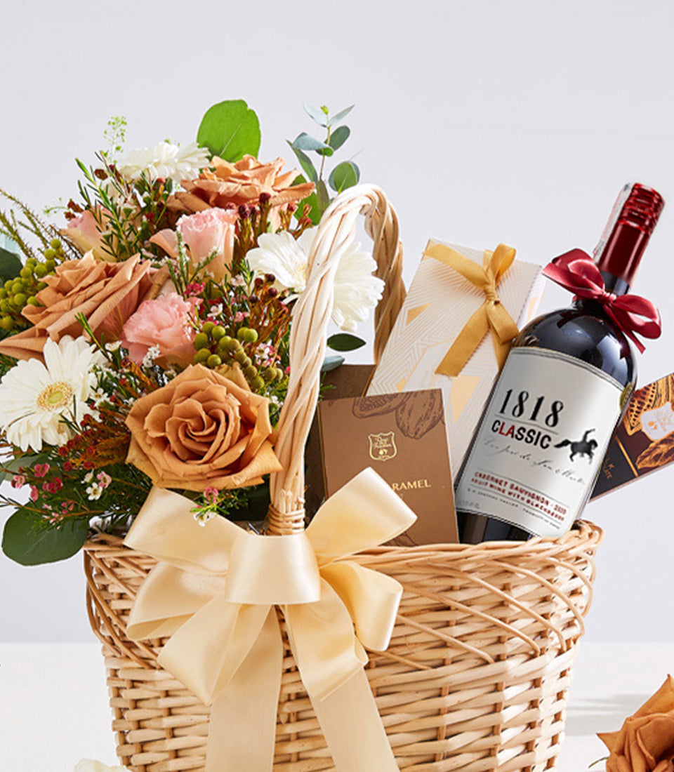 Wicker gift basket with peach and pink roses, red wine, chocolates, and gold ribbon