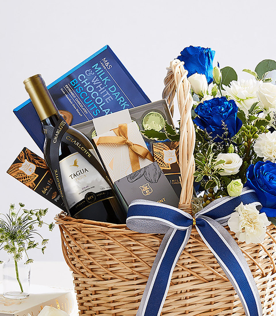 Premium wine gift basket with blue roses, white flowers, chocolate treats and gourmet snacks in woven wicker