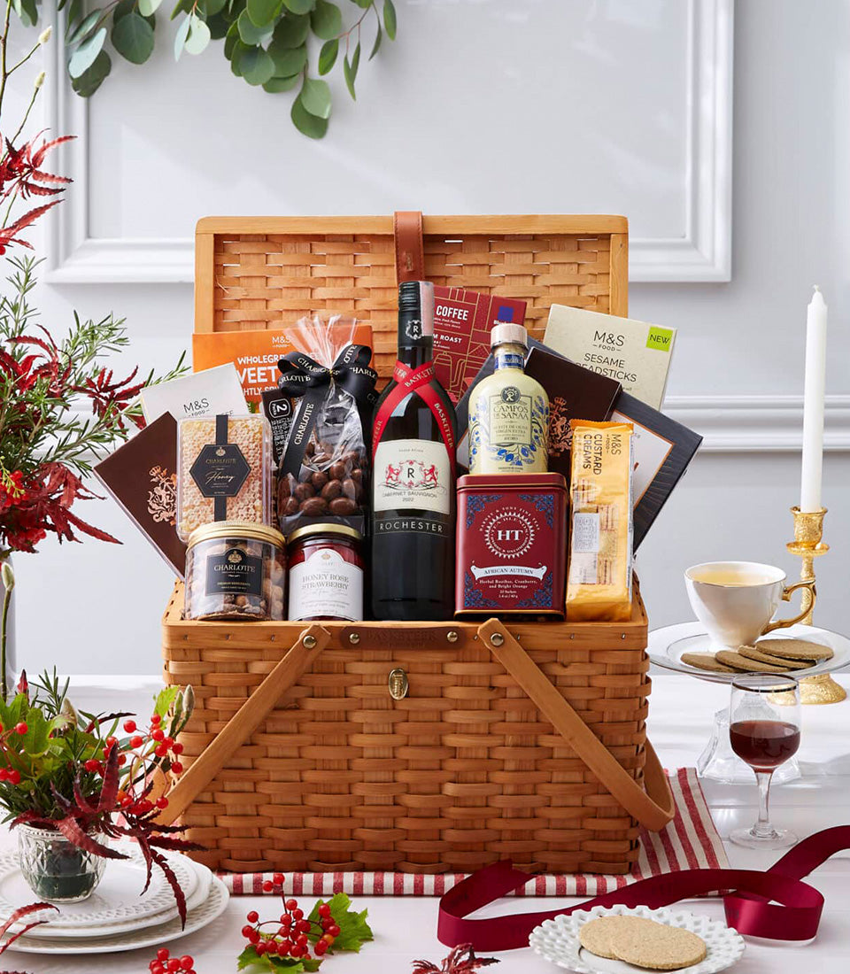 Wicker picnic basket with red wine, gourmet foods, honey, and festive red berry arrangement on white tablecloth