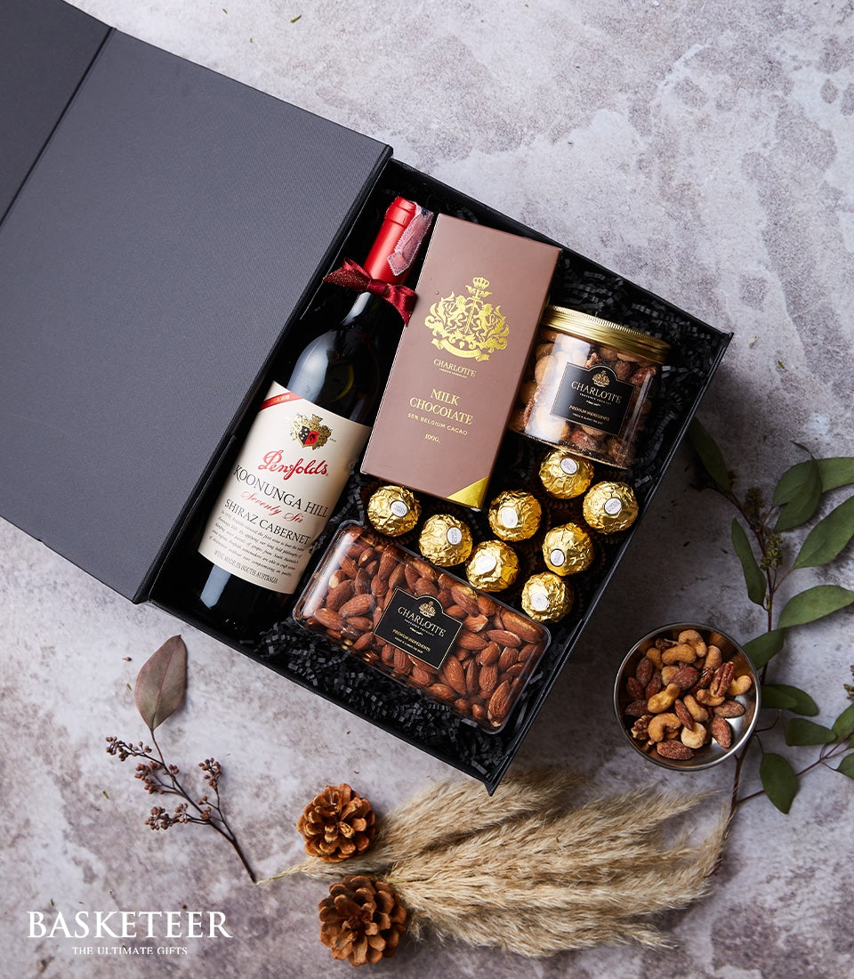 Luxury red wine and gourmet treats gift box with chocolate, nuts, and premium selections on black shredded paper