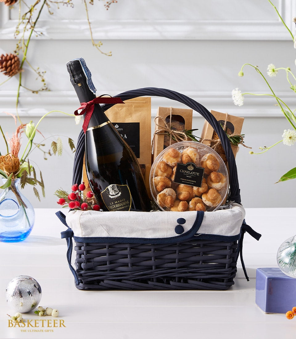 Dark blue woven gift basket with Prosecco, golden baked goods, artisan kraft pouches, and festive Christmas florals