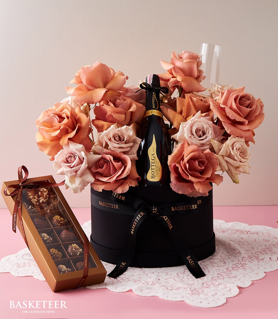 Luxury anniversary gift set with coral roses, champagne, and artisan chocolates in black hatbox