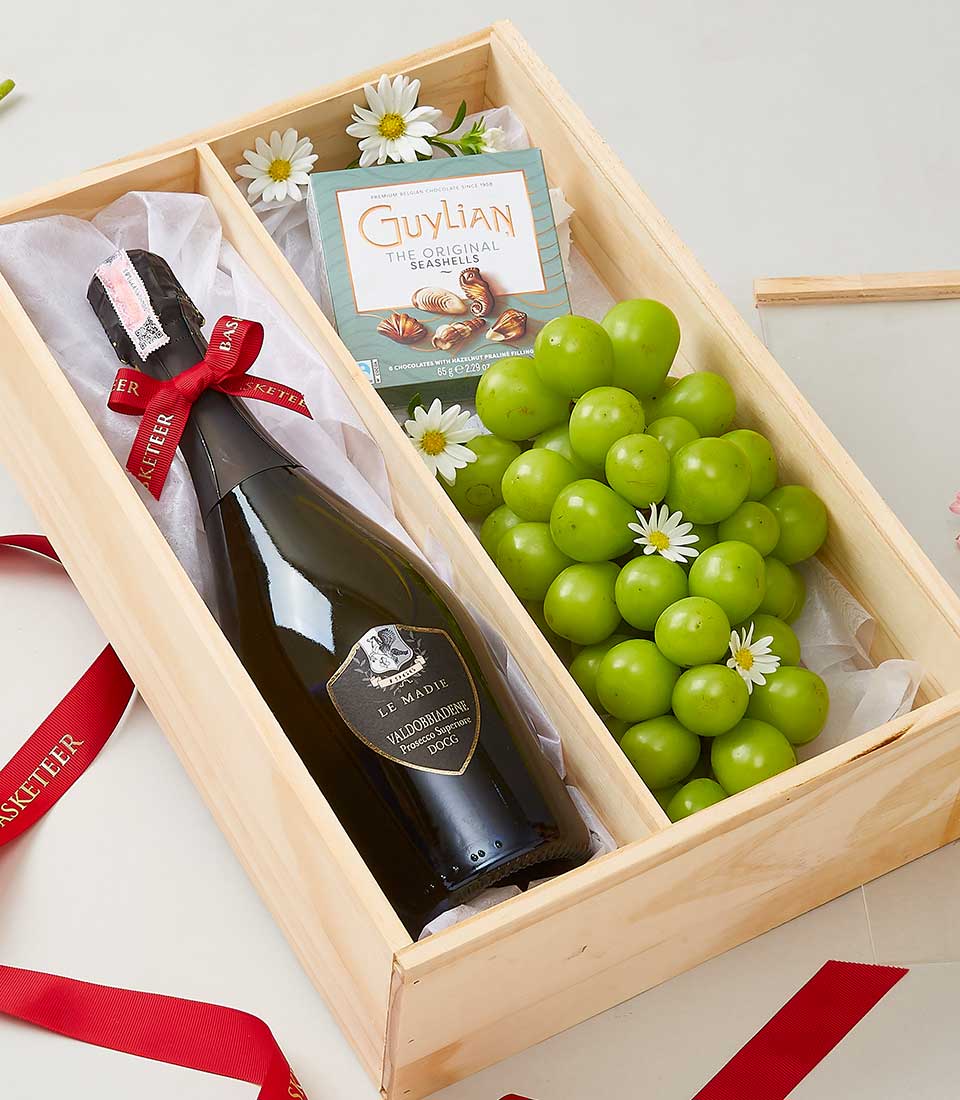 Wooden gift box with sparkling wine, seashell chocolates, and fresh green grapes tied with red ribbon