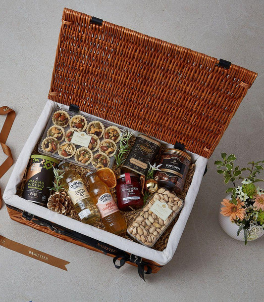 Light brown wicker picnic hamper filled with gourmet treats, artisan coffee, jam, and premium sodas, tied with black ribbon