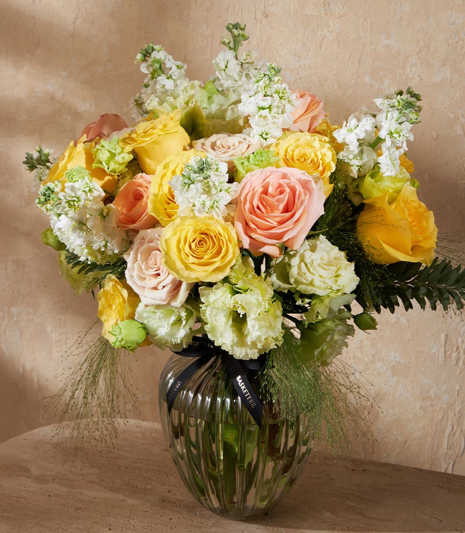 Vibrant yellow, peach and pink roses with white stock flowers in a clear ribbed glass vase