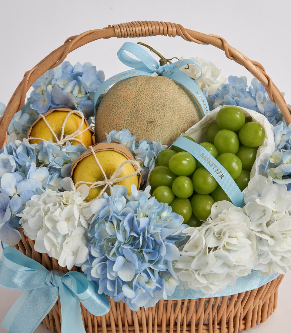 Woven wicker basket filled with fresh fruits, blue and white hydrangeas, and green ribbon bows in elegant gift presentation.