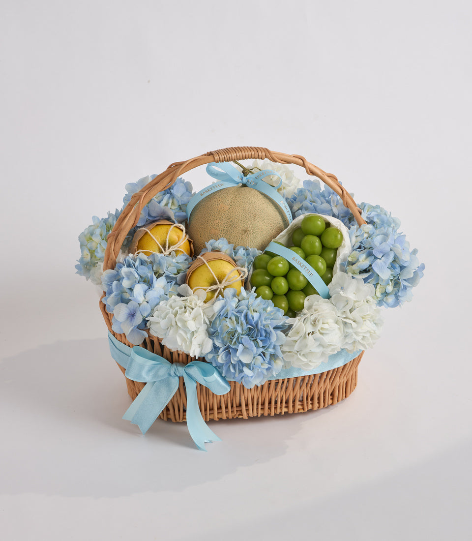 Wicker basket with blue and white hydrangeas, featuring sliced lemons, green grapes, and a tan sphere, tied with light blue ribbon.