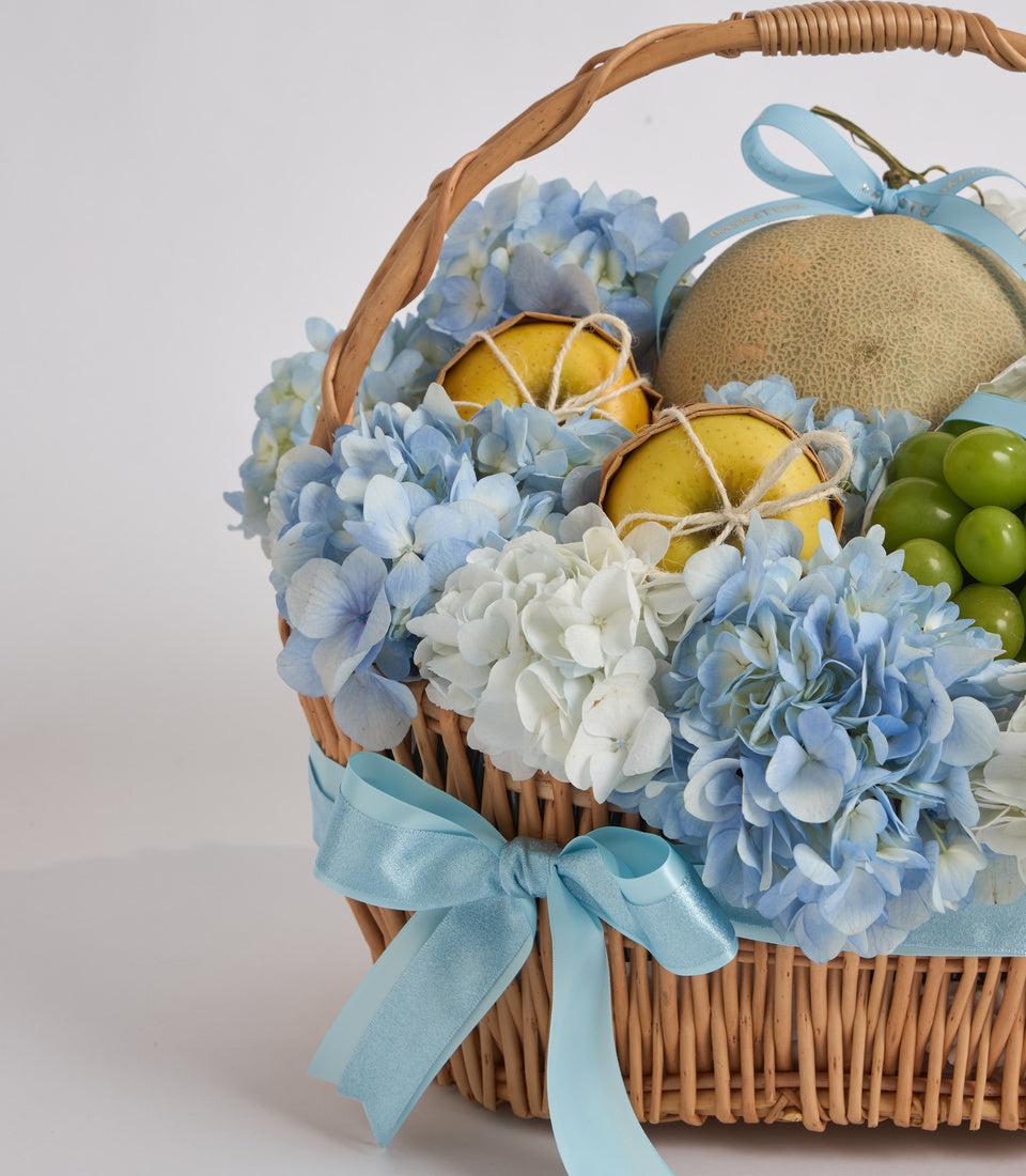 Wicker basket with blue and white hydrangeas, yellow pears with twine, green grapes, tan ornament, and blue ribbons on white background.