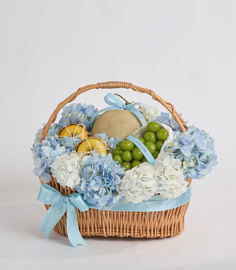 Wicker basket filled with blue and white hydrangeas, green grapes, dried lemon slices, and a melon, tied with a light blue ribbon.
