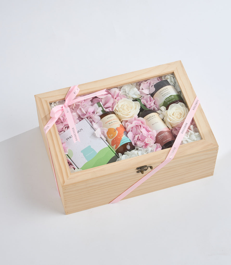 Wooden gift box with glass lid containing pink and white flowers, beauty products, and decorative items tied with pink ribbons.