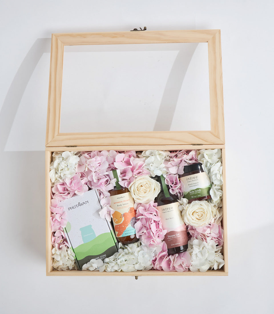 Luxury spa gift box with glass lid, natural wood frame, pink and white flowers, skincare products, and beauty items arranged on white background.