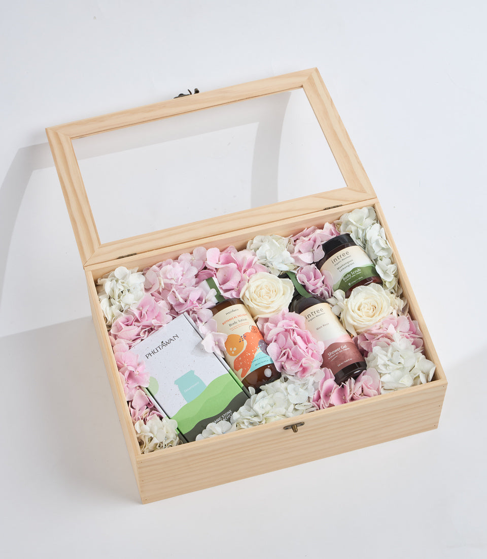Wooden gift box with glass lid containing pink and white flowers, skincare products, and a beverage bottle arranged on natural filler.
