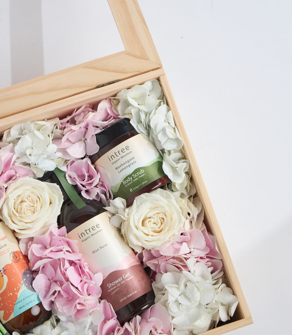 Wooden gift box filled with pink and white flowers, cream roses, and skincare products including body scrub and shower gel.