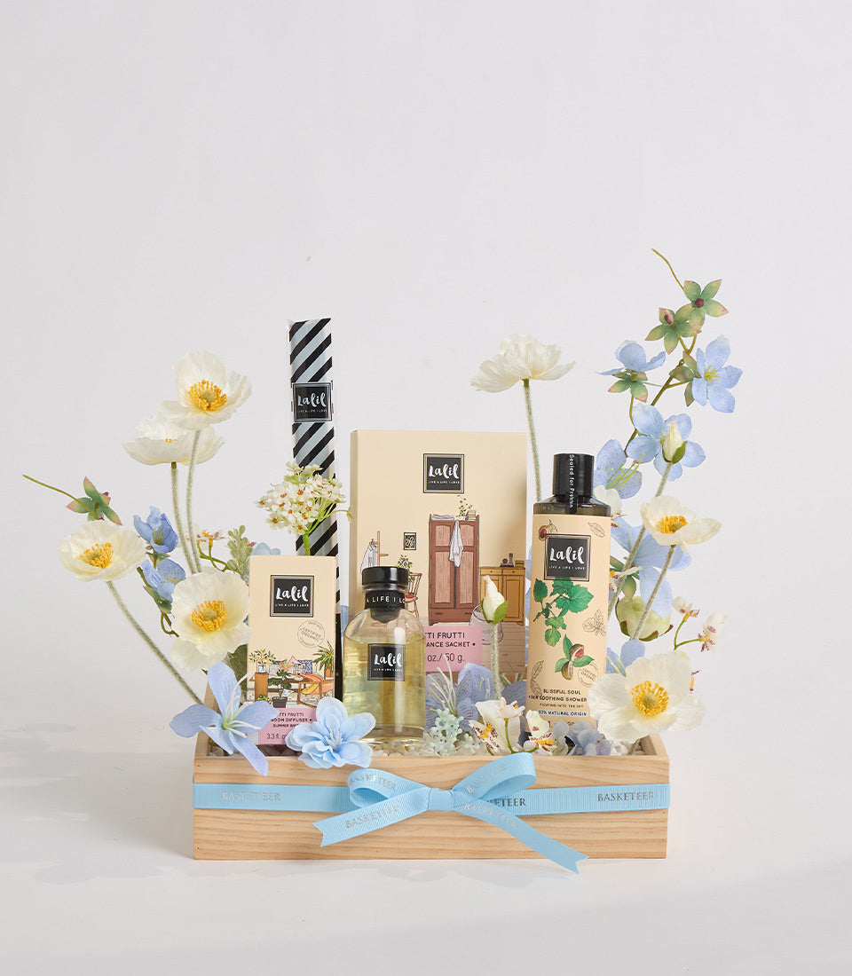Luxe gift basket with beauty products, fresh white and blue flowers, wooden box with blue ribbon bow on white background.