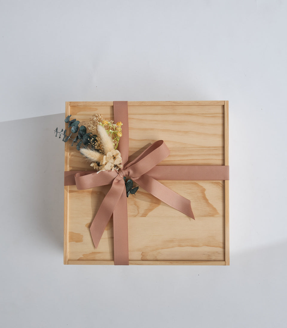Wooden gift box with mauve ribbon bow and dried flower arrangement featuring pampas grass, eucalyptus, and cream florals on light gray background.