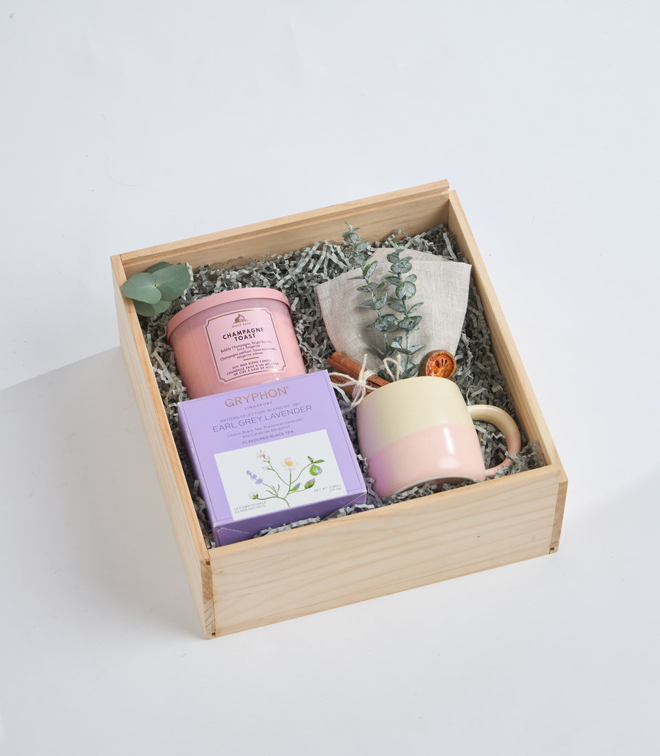 Wooden box gift set with pink candle, lavender soap, cream mug, dried eucalyptus, and shredded paper filler on white background.