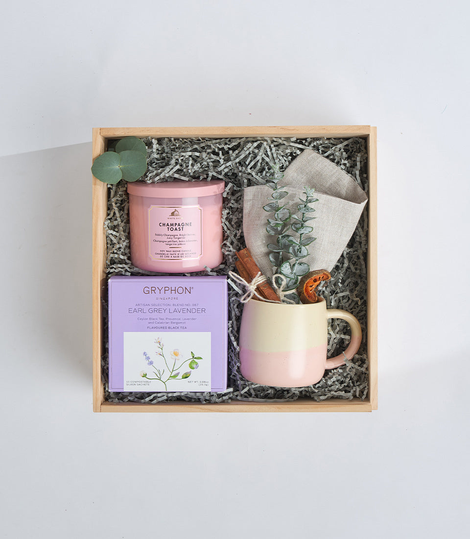 Curated gift set in wooden box featuring pink champagne candle, lavender tea, ceramic mug, linen cloth, and dried botanicals on shredded paper.