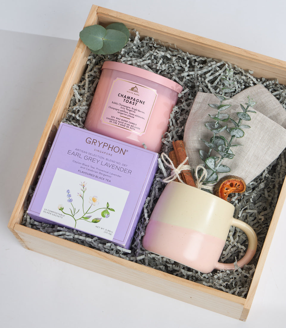 Wooden gift box containing pink Champagne Toast candle, purple Gryphon Earl Grey Lavender tea box, cream mug, herb sachet, dried citrus, cinnamon sticks, and eucalyptus leaf.