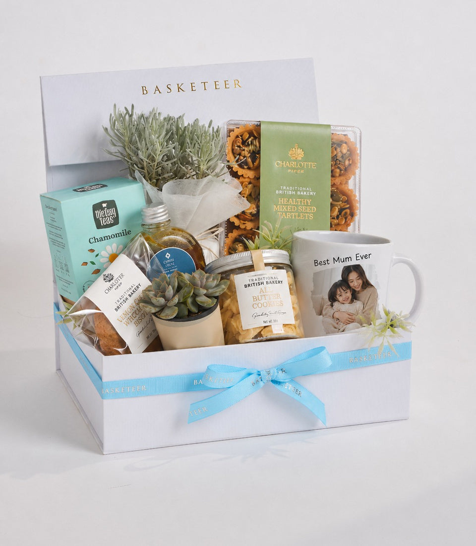 Luxurious gift basket with white box, light blue ribbon, containing plants, tea, biscuits, mug, and gourmet treats arranged elegantly.