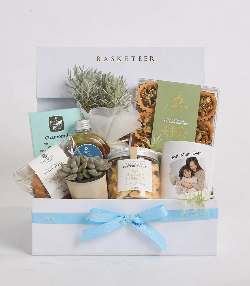 Luxury gift basket with succulents, gourmet cookies, tea, honey, and personalized photo card, tied with light blue ribbon.