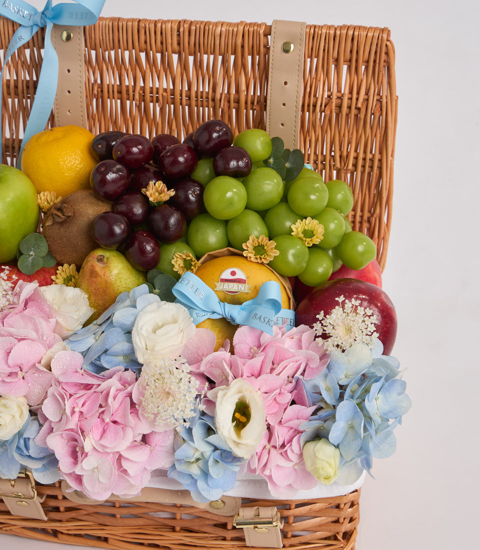 Wicker basket gift hamper with fresh fruits, pink and blue flowers, decorated with tan leather handles and light blue ribbon bow.