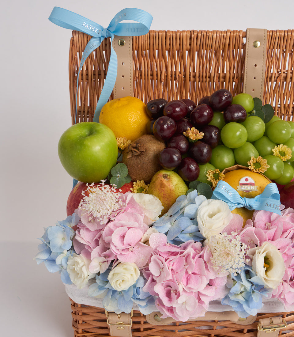 Wicker gift basket with fresh fruits, flowers, and blue ribbon bow. Contains green apples, grapes, citrus, kiwi, and pear with pink, white, and blue floral arrangement.