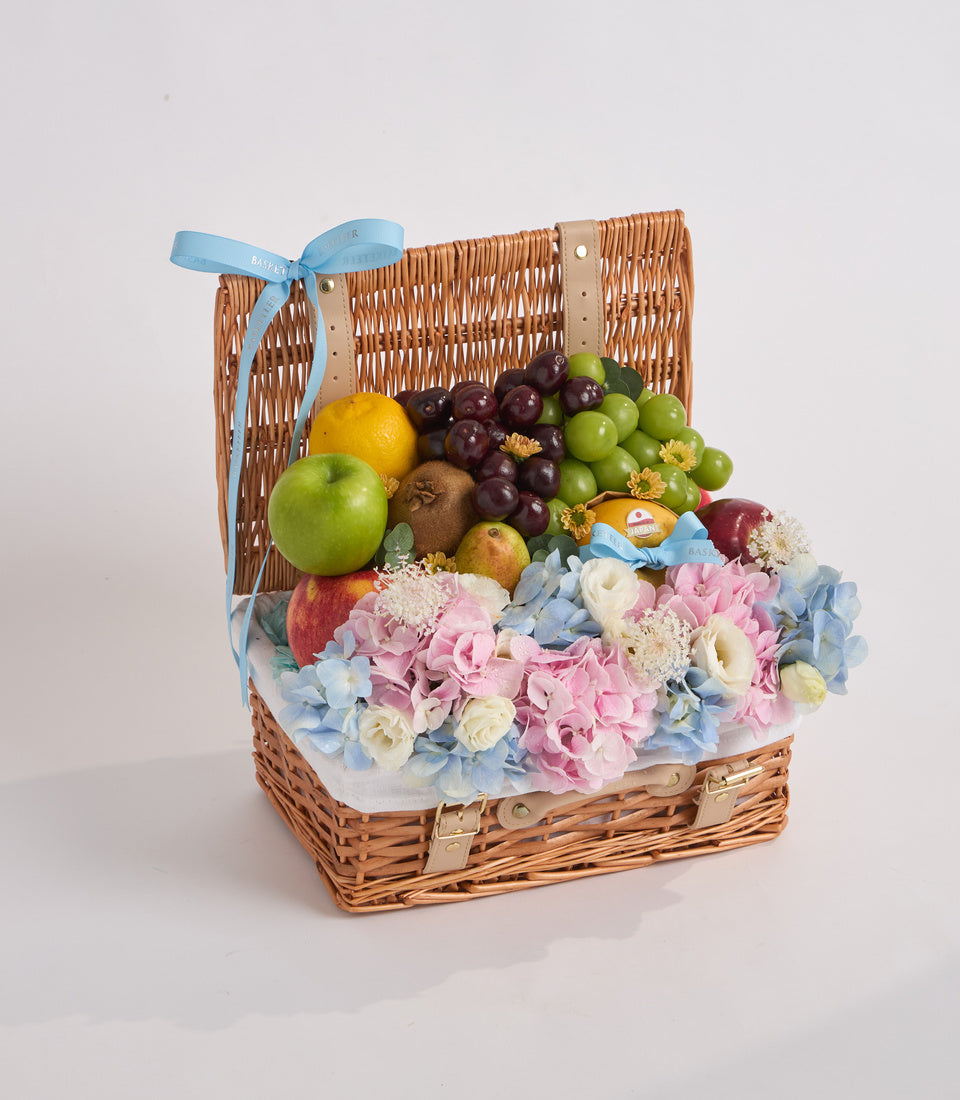 Wicker basket with blue ribbon containing fresh fruits and pink, blue, and cream flowers arranged on white tissue.