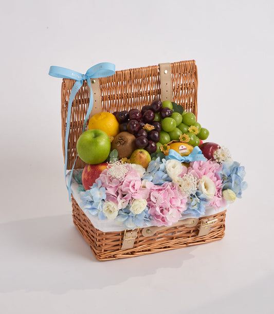 Wicker basket with blue ribbon containing fresh fruits and pink, blue, and cream flowers arranged on white tissue.