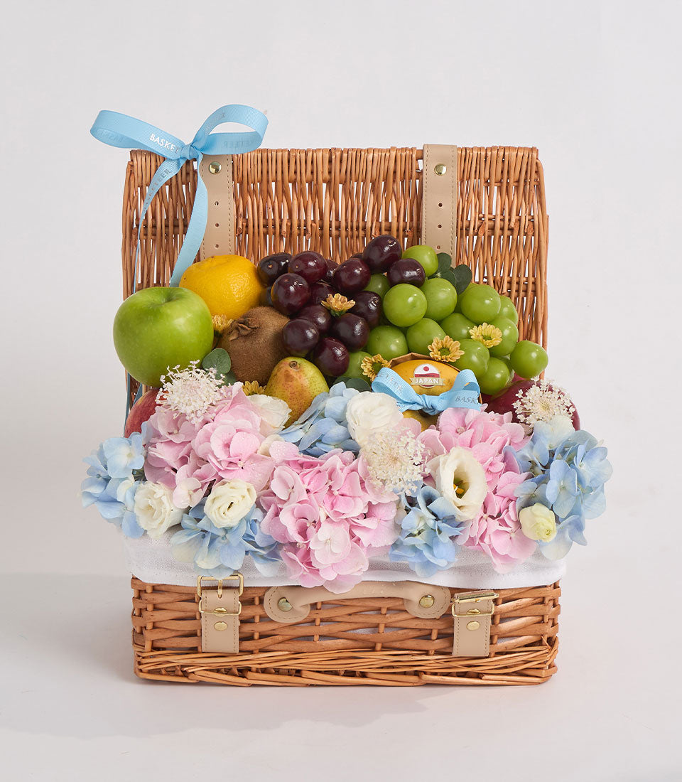 Wicker picnic basket with leather handles filled with fresh fruits and colorful flowers in pink, blue, and white tones with blue ribbon.