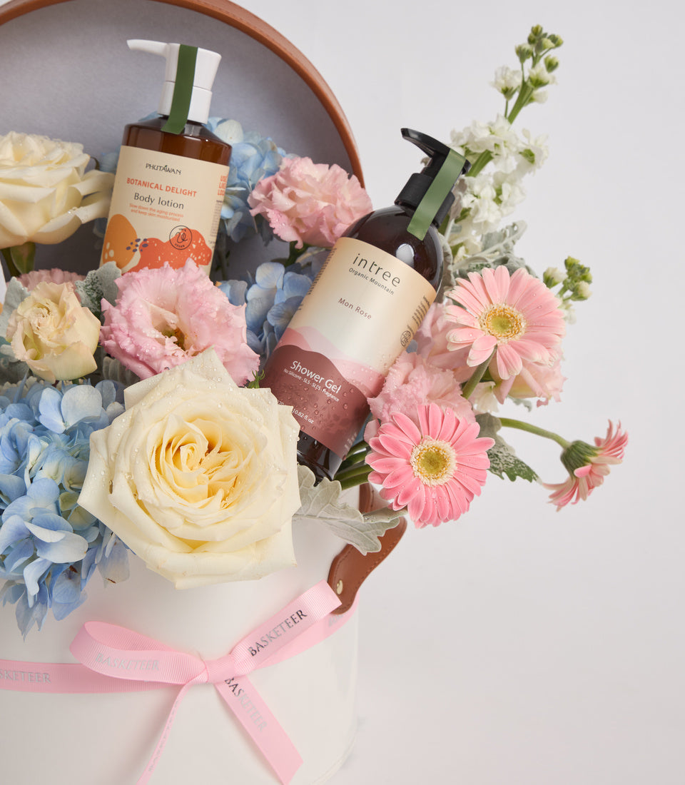 Skincare gift set with two brown bottles nestled in a ceramic bowl surrounded by fresh flowers in pink, yellow, and blue tones, tied with pink ribbon.