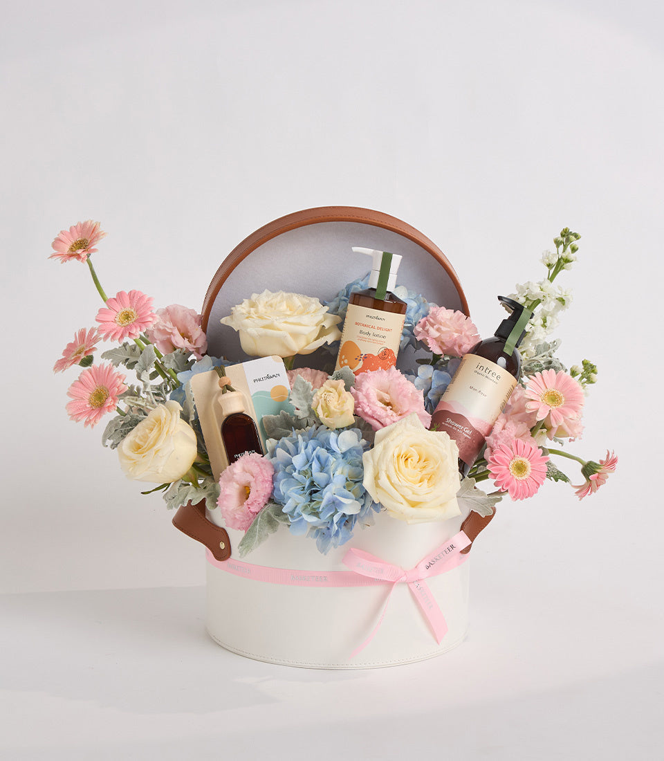 Luxury beauty gift basket with pink gerberas, cream roses, blue hydrangeas, skincare bottles, wooden handle, white box, pink ribbon bow.