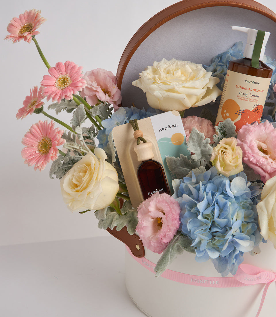 Luxurious gift basket with pink gerberas, cream roses, blue hydrangeas, Phutawan skincare products, and wooden handles with pink ribbon.
