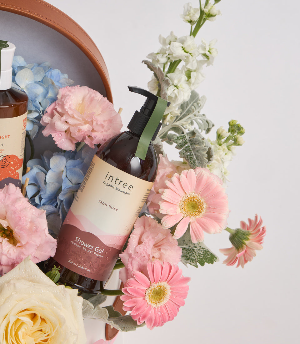 Intree Mon Rose Shower Gel surrounded by pink gerberas, carnations, white flowers, and blue hydrangeas in a terracotta bowl against white background.