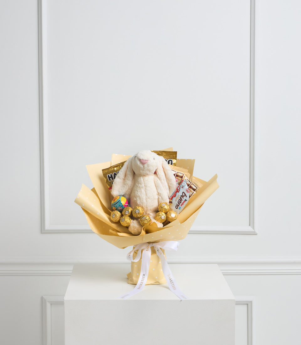 Luxe gift bouquet with cream bunny plush, gold Ferrero chocolates, treats, and gold cellophane wrap with lavender ribbon on white pedestal.