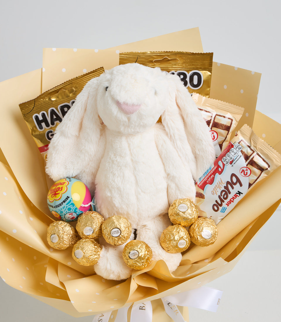 White fluffy bunny plush toy surrounded by gold foil chocolates, candies, and snacks in cream and gold polka dot wrapping paper bouquet.
