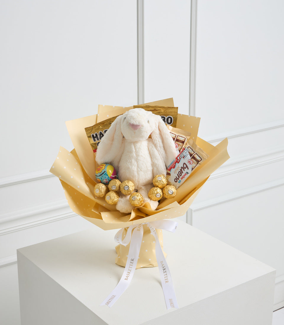 Luxury gift bouquet with white plush bunny rabbit, gold-wrapped chocolates, snacks, and yellow tissue paper wrapping with white ribbon on white pedestal.