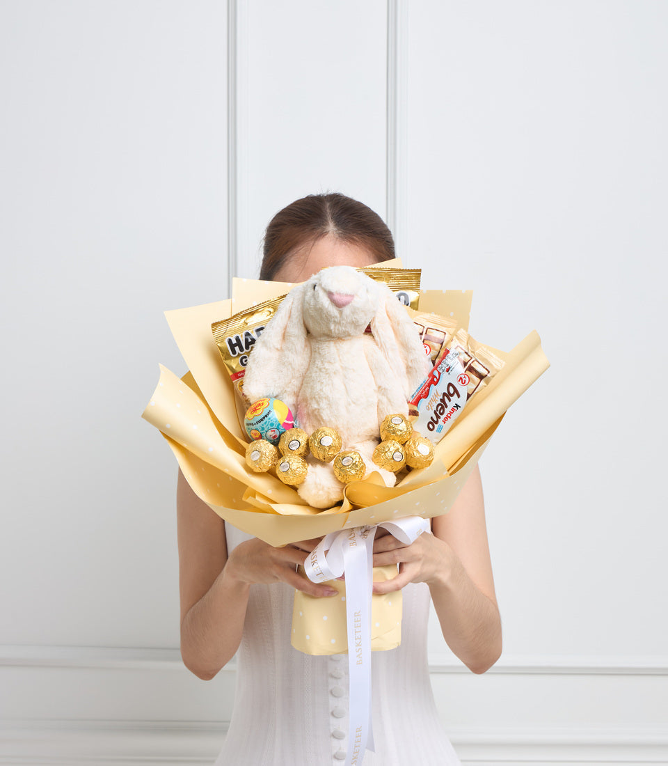 Woman holding luxury gift bouquet with cream plush bunny rabbit, gold wrapped chocolates, snacks, and golden tissue paper wrapping with white ribbon.