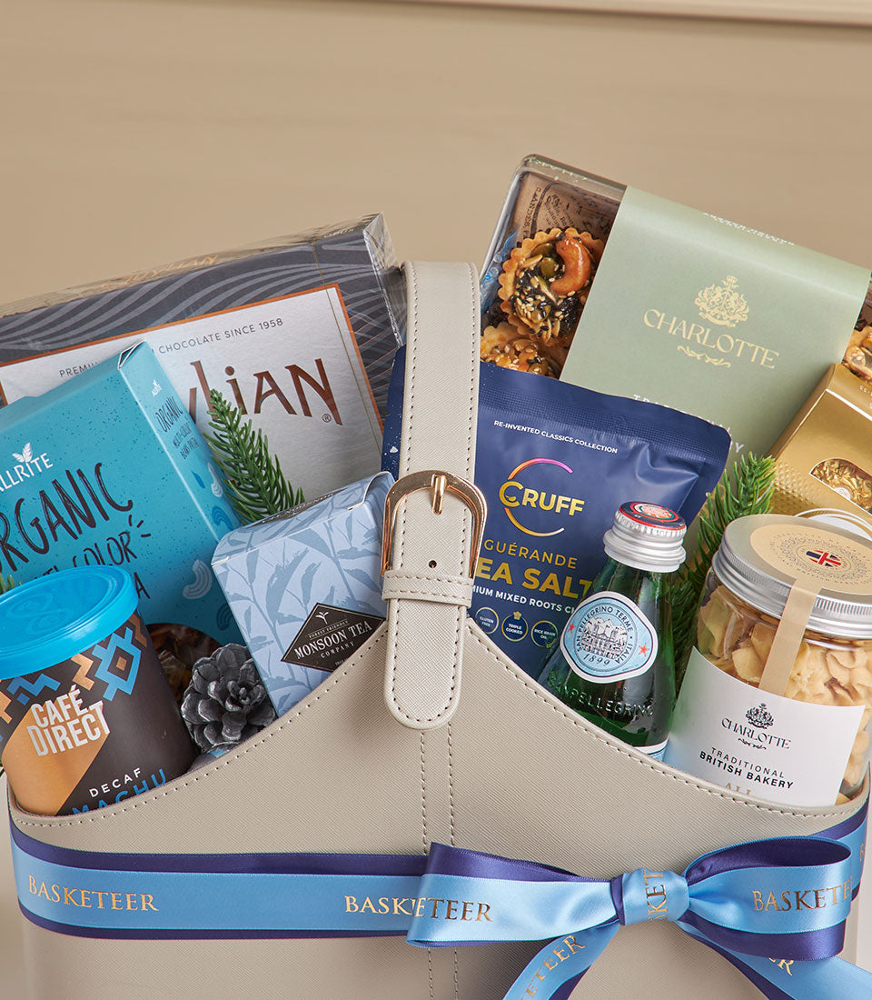Beige leather gourmet tea and coffee gift basket with gold buckle, blue ribbon, artisan chocolates, and premium treats for