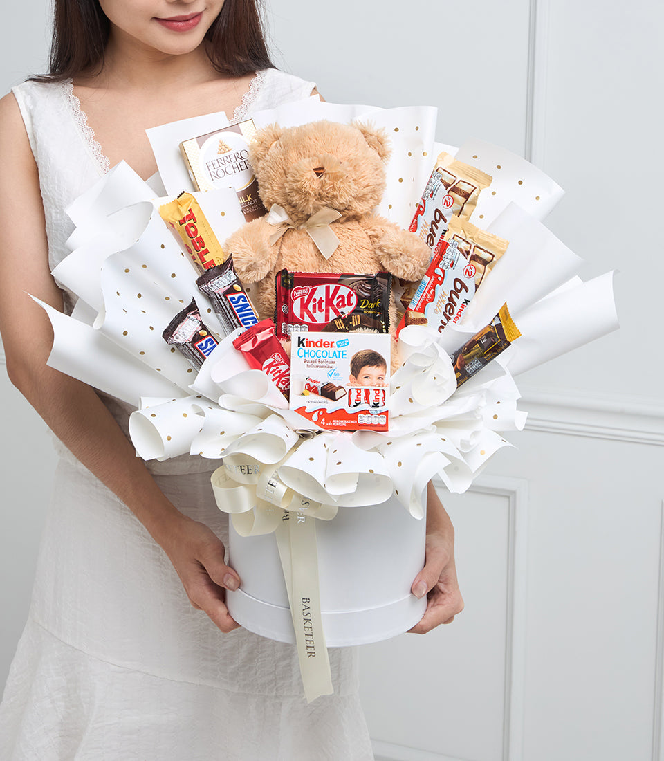 Woman in white dress holding decorative chocolate gift bouquet with teddy bear, white paper wrapping, gold dots, and assorted chocolates including Ferrero Rocher and Kinder.