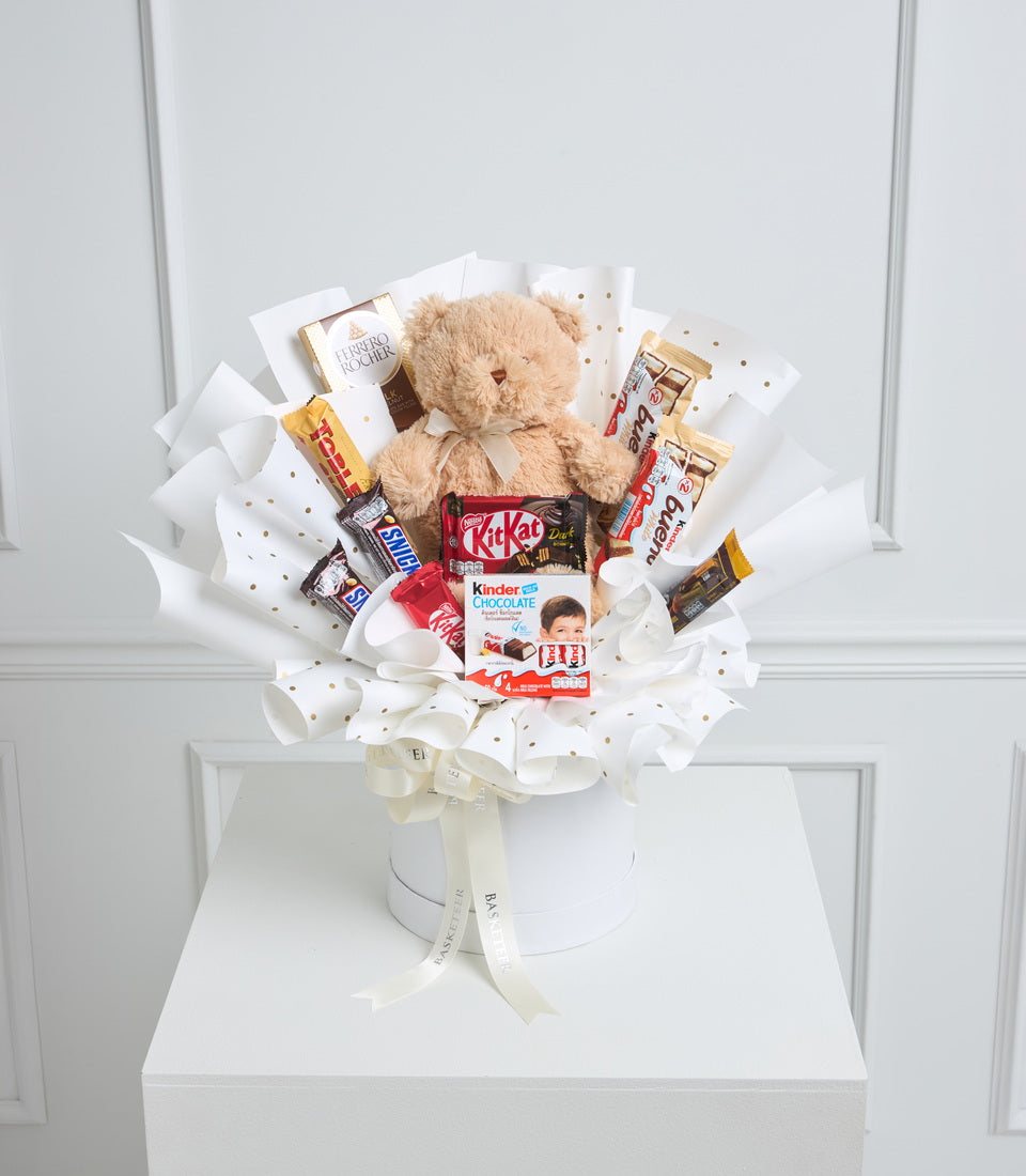 Teddy bear surrounded by chocolate and candy treats in white box with gold dots and cream ribbon bow.