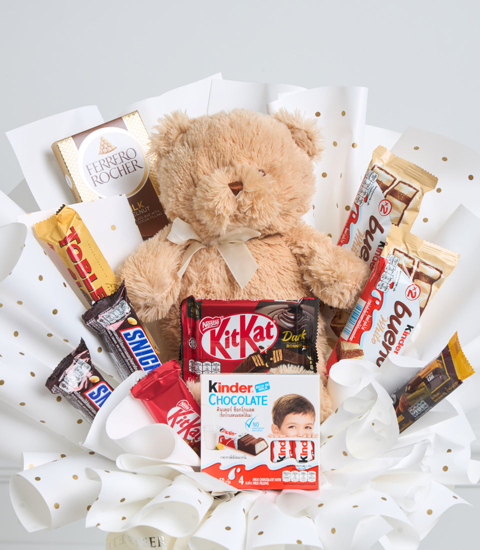 Cuddly teddy bear surrounded by assorted chocolates and sweets including Ferrero Rocher, Kit Kat, Kinder, and Snickers on white tissue paper with gold polka dots.