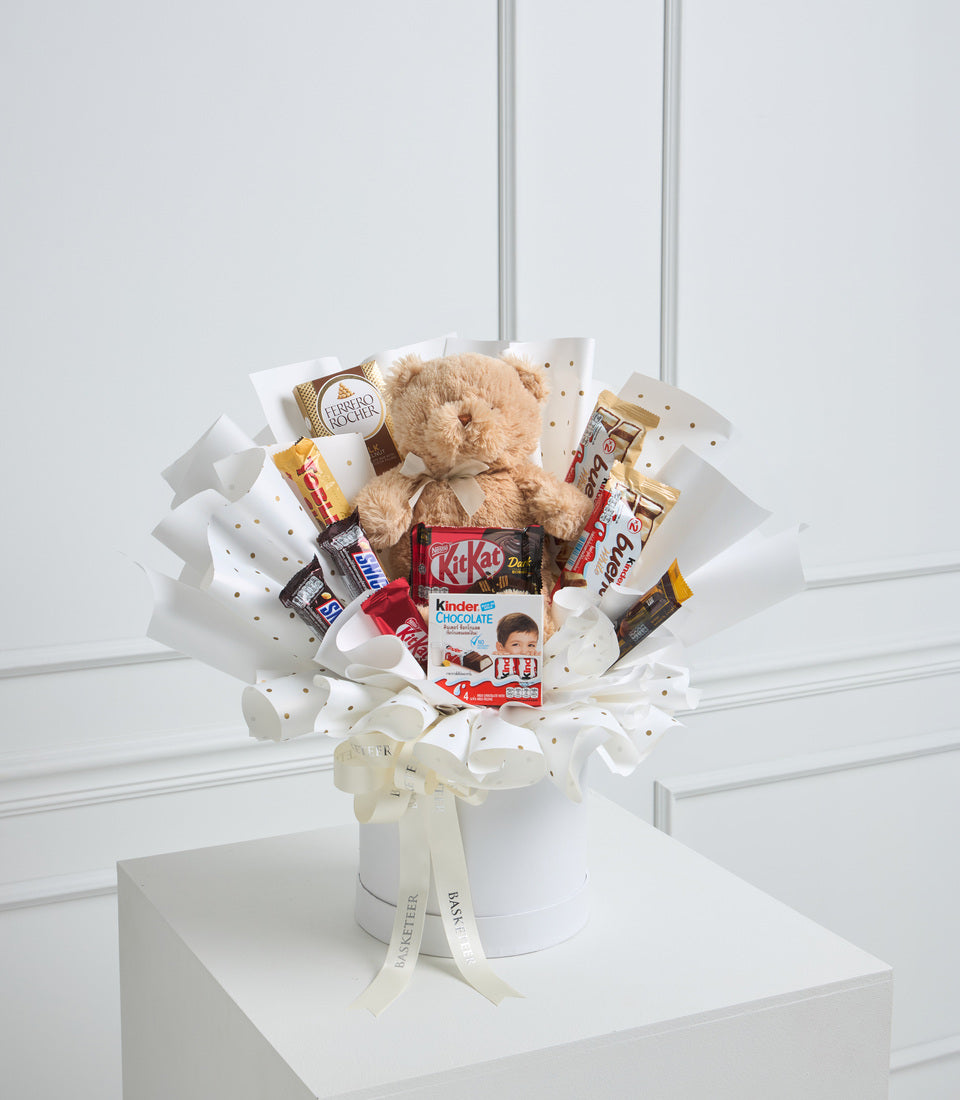 Teddy bear surrounded by chocolate and candy treats in white gift box with gold polka dots, cream ribbon bow, luxury gift arrangement.