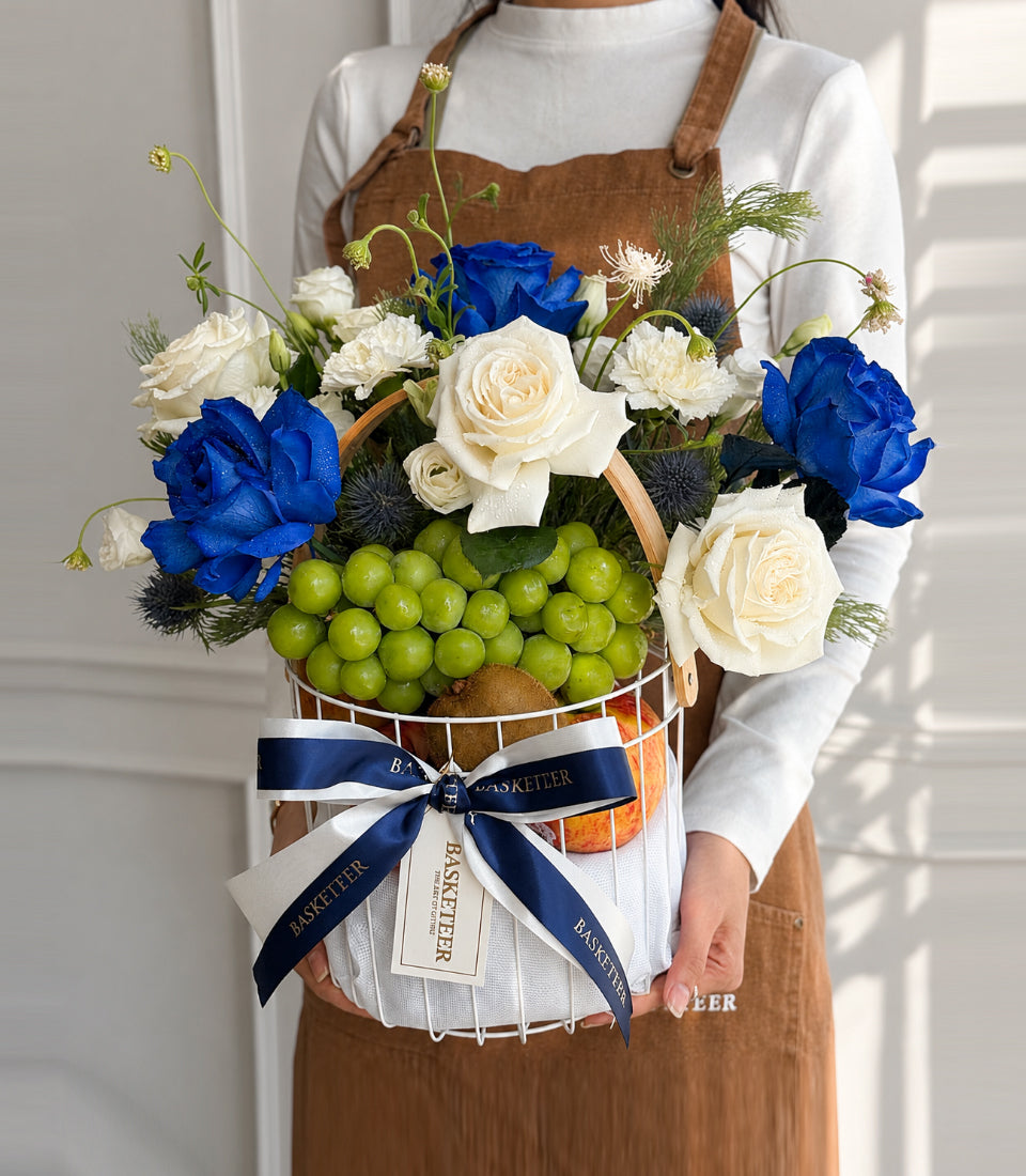 Florist holding luxury basket arrangement with blue roses, cream roses, green grapes, dark thistle, and navy ribbon bow on white background.